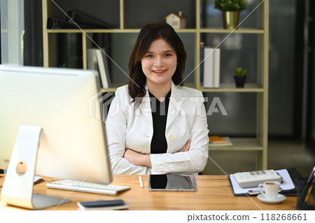 Attractive young businesswoman sitting at her workplace with arms crossed and smiling 118268661