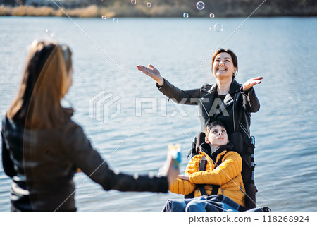 Mother and children enjoying outdoor time by the lake, inclusive family activities, child in wheelchair, sibling blowing bubbles, disability inclusion, family bonding. 118268924
