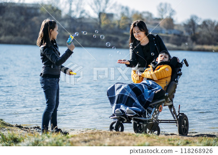 Mother and children enjoying outdoor time by the lake, inclusive family activities, child in wheelchair, sibling blowing bubbles, disability inclusion, family bonding. 118268926