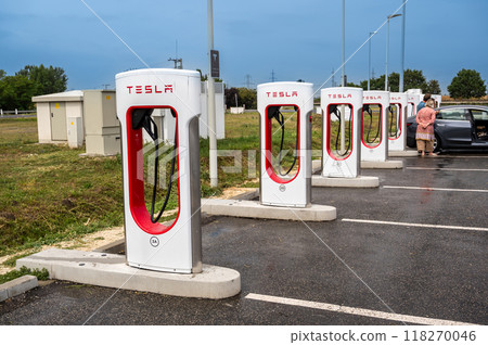Highway to Budapest, Hungary. August 24, 2022. Tesla fast charging stations in a rest area. In the background a car is refueling. The owners are next to the car. Highway to Budapest, Hungary. August 24, 2022. Tesla fast charging stations in a rest area. In the background a car is refueling. The owners are next to the car. 118270046