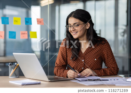 Happy woman financier doing paperwork inside office, latin american smiling pleased preparing financial report, accountant recording data and filling forms sitting at table with documents and laptop. Happy woman financier doing paperwork inside office, latin american smiling pleased preparing financial report, accountant recording data and filling forms sitting at table with documents and laptop. 118271289