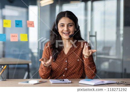 Webcam view, video call online meeting, woman with headset phone smiling and looking at camera, talking remotely with clients and colleagues, business woman at workplace inside office. Webcam view, video call online meeting, woman with headset phone smiling and looking at camera, talking remotely with clients and colleagues, business woman at workplace inside office. 118271292