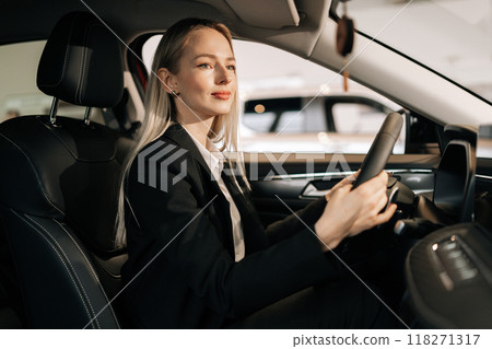 Attractive young blonde woman customer sitting on driver seat inside car, choosing automobile test drive in dealership. Smiling female buyer touching steering wheel dreaming buying new transport. Attractive young blonde woman customer sitting on driver seat inside car, choosing automobile test drive in dealership. Smiling female buyer touching steering wheel dreaming buying new transport. 118271317