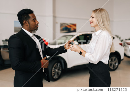Side view of black salesman and female buyer shaking hands after successful deal in dealership center. African salesperson giving car keys to client, greeting with purchase in auto showroom. 118271323