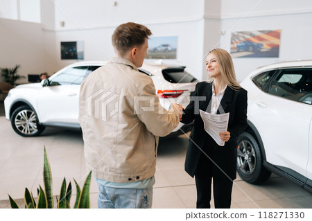 Rear view of happy male car owner getting key from smiling female auto dealer at dealership. Handsome young male taking key to bought car and sealing deal with handshake in auto showroom 118271330