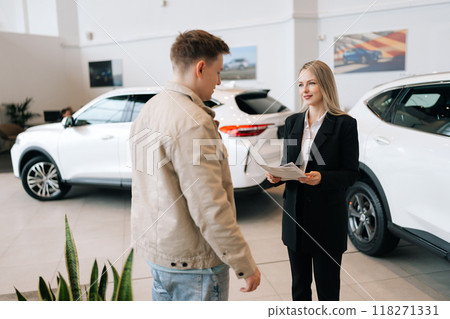 Attractive professional female car dealer in suit holding paper document, explaining buying new car to young man at modern showroom. Concept of choosing and buying new auto in dealership office. Attractive professional female car dealer in suit holding paper document, explaining buying new car to young man at modern showroom. Concept of choosing and buying new auto in dealership office. 118271331