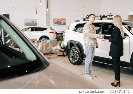 Remote wide shot of happy male car owner getting key from smiling female auto dealer at dealership. Handsome young male taking key to bought car and sealing deal with handshake in auto showroom Remote wide shot of happy male car owner getting key from smiling female auto dealer at dealership. Handsome young male taking key to bought car and sealing deal with handshake in auto showroom 118271342