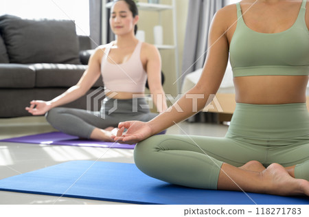 Two women doing yoga meditating exercise at home, sitting in Easy seat pose with mudra in  bedroom. Working out wearing sportive top and leggings. Relaxation and Healthcare concept. 118271783