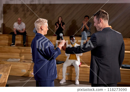 Rear view on man giving microphone to adult woman in formal wear for speaking Rear view on man giving microphone to adult woman in formal wear for speaking 118273090