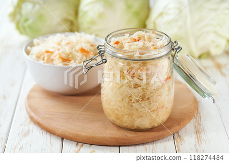 Open glass jar of fermented white cabbage with carrot on a white kitchen table. Open glass jar of fermented white cabbage with carrot on a white kitchen table. 118274484