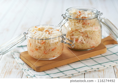 Open glass jar of fermented white cabbage with carrot on a white kitchen table. 118274492