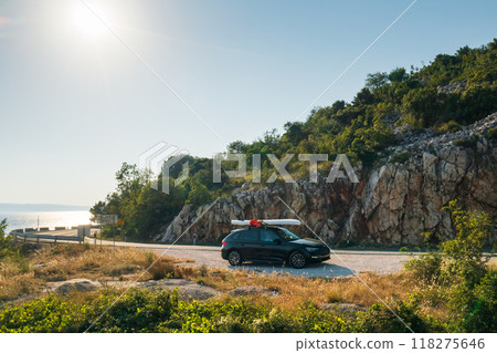 Black car with a paddleboard firmly strapped to its roof, parked on a cliffside road overlooking the deep blue sea water 118275646