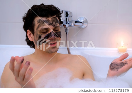 Man with charcoal face mask lying in bath with foam candle meditating at home. 118278179