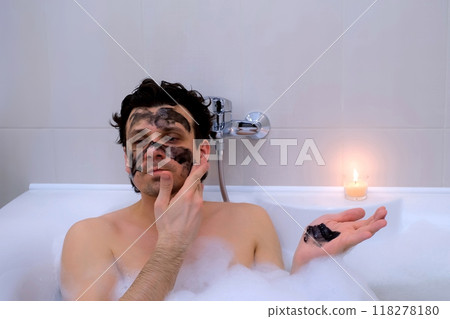 Man applying charcoal face mask lying in bath with foam and candle at home. 118278180