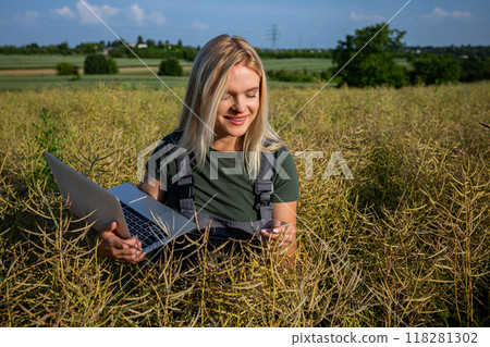 A smiling woman farmer checking the quality of rapeseed, standing at the rapeseed field and holding a laptop 118281302
