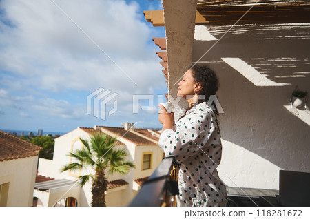 Woman enjoying morning coffee on sunny Mediterranean balcony with peaceful view of rooftops and palm trees. 118281672