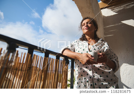 Woman enjoying morning coffee on sunny balcony in polka dot shirt, feeling relaxed and thoughtful Woman enjoying morning coffee on sunny balcony in polka dot shirt, feeling relaxed and thoughtful 118281675