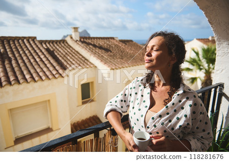 Woman enjoying a sunny morning on a balcony, feeling relaxed and peaceful while holding a cup of coffee, with charming Mediterranean architecture in the background. Woman enjoying a sunny morning on a balcony, feeling relaxed and peaceful while holding a cup of coffee, with charming Mediterranean architecture in the background. 118281676