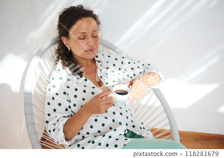 Woman relaxing with a cup of coffee in a sunlit room, wearing a white polka dot shirt, enjoying quiet moments and calm atmosphere Woman relaxing with a cup of coffee in a sunlit room, wearing a white polka dot shirt, enjoying quiet moments and calm atmosphere 118281679