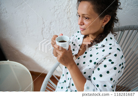 Relaxed woman enjoying a cup of coffee in a cozy chair by a textured wall, wearing a white shirt with green polka dots 118281687
