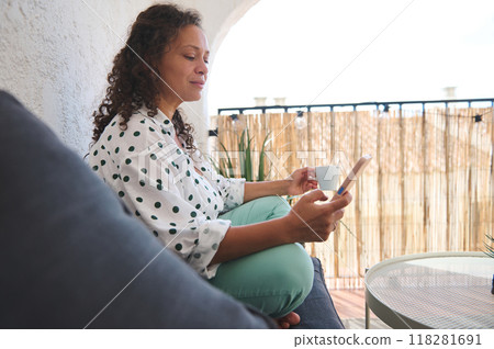 Relaxed woman enjoying a coffee while browsing on mobile phone in cozy balcony setting with natural light Relaxed woman enjoying a coffee while browsing on mobile phone in cozy balcony setting with natural light 118281691