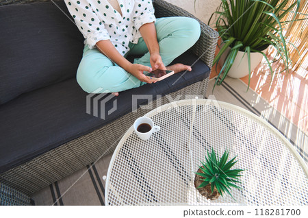 Woman relaxing on a patio sofa holding a smartphone with coffee and a plant on a wicker table for a peaceful outdoor setting 118281700