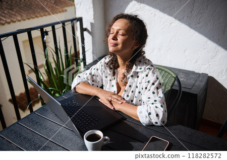 Woman enjoying a peaceful moment on balcony with laptop and coffee, absorbing the sunlight, wearing a polka dot shirt Woman enjoying a peaceful moment on balcony with laptop and coffee, absorbing the sunlight, wearing a polka dot shirt 118282572