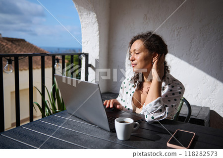 Woman enjoying remote work on a sunny balcony with a laptop, cup of coffee, and smartphone in a serene setting 118282573