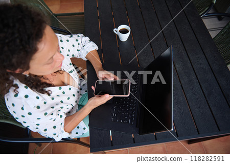 Woman using smartphone and laptop while working remotely on a patio with a cup of coffee, wearing a polka dot shirt. Woman using smartphone and laptop while working remotely on a patio with a cup of coffee, wearing a polka dot shirt. 118282591
