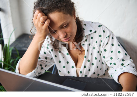 Woman in polka dot shirt appears stressed while working at a laptop in a modern home office setting 118282597