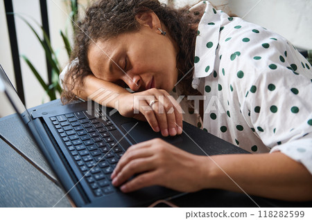 Woman sleeping at a desk on laptop, exhausted from work, wearing a polka dot shirt, representing fatigue and burnout in the workplace. 118282599