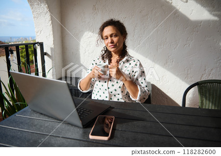 Relaxed woman enjoying coffee break while working on a laptop from a sunlit balcony with a beautiful view 118282600