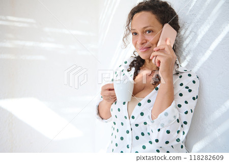 Relaxed woman enjoying a coffee break at home while having a pleasant phone conversation on a sunny day Relaxed woman enjoying a coffee break at home while having a pleasant phone conversation on a sunny day 118282609