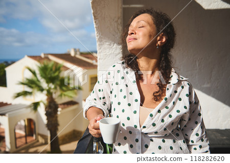 Woman enjoying morning sunlight on a sunny balcony while sipping coffee, wearing a polka dot shirt, and embracing a moment of relaxation. Woman enjoying morning sunlight on a sunny balcony while sipping coffee, wearing a polka dot shirt, and embracing a moment of relaxation. 118282620