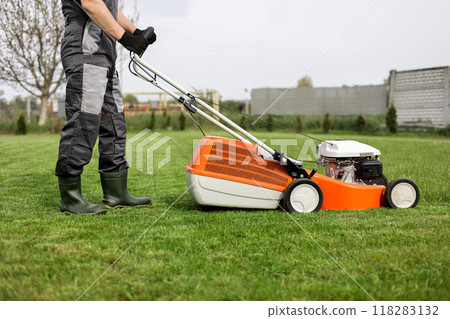Professional gardener in protective apparel is mowing green grass lawn using modern gasoline cordless lawnmower at backyard. Seasonal landscaping design work. Blooming trees on background Professional gardener in protective apparel is mowing green grass lawn using modern gasoline cordless lawnmower at backyard. Seasonal landscaping design work. Blooming trees on background 118283132