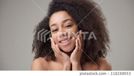 Beauty and healthcare concept - beautiful African American woman with curly afro hairstyle and clean, healthy skin touching face with hand, doing facial massage, posing and looking at camera 118289053
