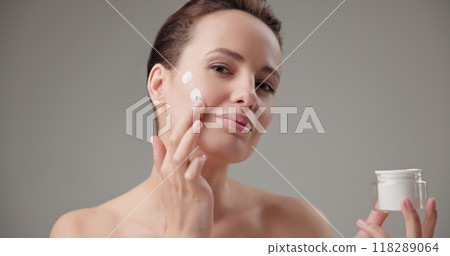 Beauty, health, cosmetics, anti-aging therapy and skin care concept - young beautiful brunette Caucasian woman holds a jar of moisturizer in her hands and applies the cream to her face skin Beauty, health, cosmetics, anti-aging therapy and skin care concept - young beautiful brunette Caucasian woman holds a jar of moisturizer in her hands and applies the cream to her face skin 118289064