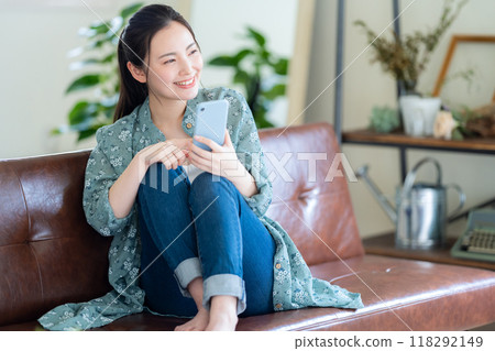 一位年輕女子在客廳一邊放鬆一邊看著智慧型手機 118292149