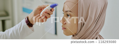 Profile view of woman wearing hijab receiving temperature check using infrared thermometer device in medical setting without natural light 118293080