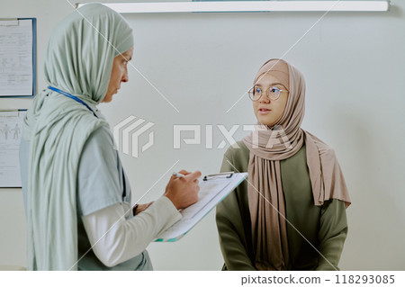 Healthcare professional in headscarf talking to patient while taking notes. Medical consultation focusing on understanding and discussing patients health needs 118293085
