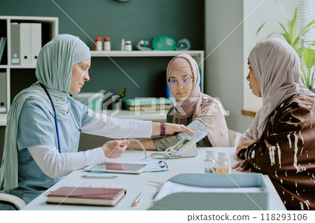 Three Muslim women with hijabs discussing medical procedures in healthcare office with medical equipment on desk and shelves in background 118293106