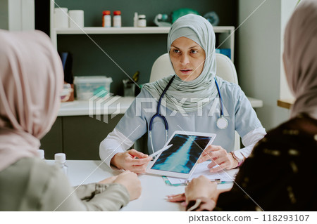 Medical professional in headscarf consulting patients while holding tablet in examination room. Clinical setting includes modern equipment and shelves storing medical supplies Medical professional in headscarf consulting patients while holding tablet in examination room. Clinical setting includes modern equipment and shelves storing medical supplies 118293107