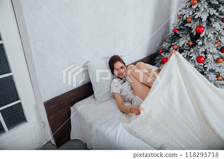 Woman in pajamas on bed in bedroom woke up by Christmas tree with gifts Woman in pajamas on bed in bedroom woke up by Christmas tree with gifts 118293718