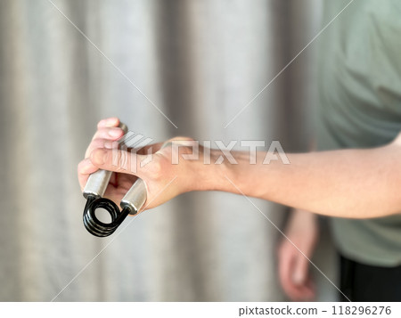 Close up of hand man holding black and silver hand expander. Focus on hand holding pen with blurred background. Concept of fitness, exercise and healthy lifestyle at home. Close up of hand man holding black and silver hand expander. Focus on hand holding pen with blurred background. Concept of fitness, exercise and healthy lifestyle at home. 118296276