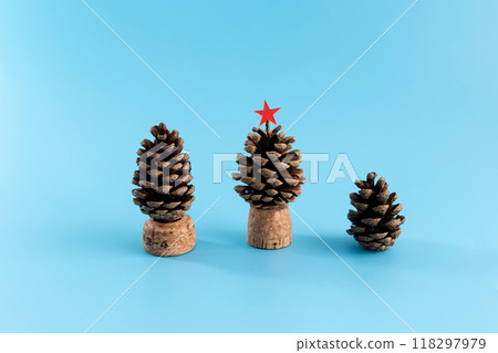 Three pine cones of different sizes on blue surface, mounted on cork bases, resembling tiny trees. Large pine cone with red star top, showcasing festive DIY craft with natural materials 118297979