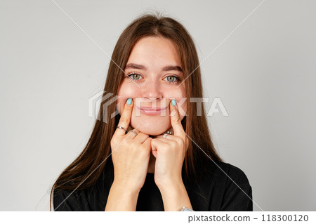 Young Woman Smiling With Hands on Cheeks Against a Gray Background 118300120