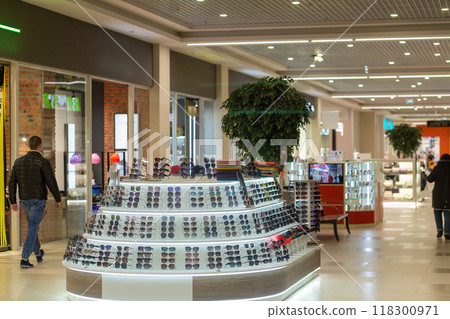 An elegant sunglasses display occupies the center of a busy mall, surrounded by shoppers and retail stores. An elegant sunglasses display occupies the center of a busy mall, surrounded by shoppers and retail stores. 118300971