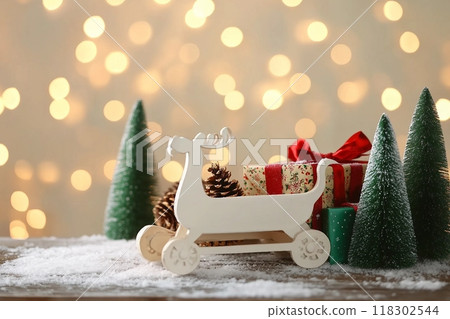 Small white sleigh, decorative Christmas trees and gift boxes on wooden table against blurred lights Small white sleigh, decorative Christmas trees and gift boxes on wooden table against blurred lights 118302544