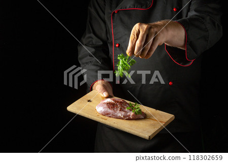 Chef sprinkles salt on meat steak with hand before grilling. Hotel or restaurant service concept on black background with place for advertising. Cooking meat in kitchen 118302659