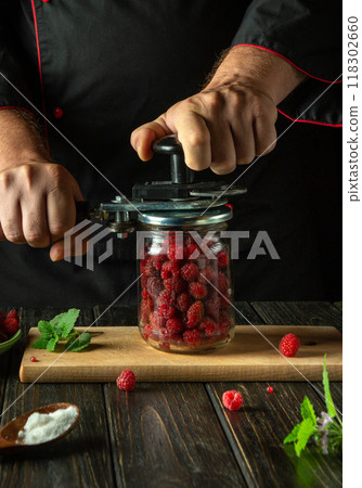 Chef seals ripe raspberries in a jar with a manual seaming key. Concept of making canned sweet drink or jam with Rubus idaeus on the kitchen table Chef seals ripe raspberries in a jar with a manual seaming key. Concept of making canned sweet drink or jam with Rubus idaeus on the kitchen table 118302660